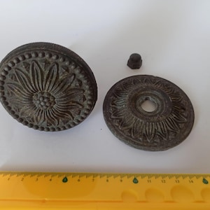 May include: Antique bronze-coloured hardware components, including a large, round piece with a floral design, a smaller circular piece, and a small nut. The floral design is detailed with a central flower and surrounding petals. A yellow ruler is in the foreground.