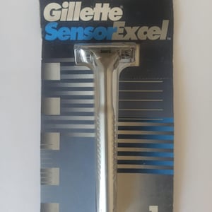 May include: A Gillette SensorExcel razor in its original packaging. The packaging is dark blue with silver and blue stripes. The razor is silver with a textured handle. The text "Gillette SensorExcel" is printed on the packaging.