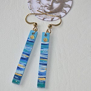 May include: A pair of rectangular earrings with a blue, white, and gold painted design. The earrings hang from gold-coloured hooks. The design resembles a seascape. A circular image of a face is visible in the background.