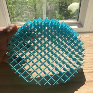 May include: A round, light blue plastic grid with a crosshatch pattern. The grid is held up in front of a window, with sunlight casting shadows on the wooden surface below. The grid is made of thin, interconnected plastic strips.