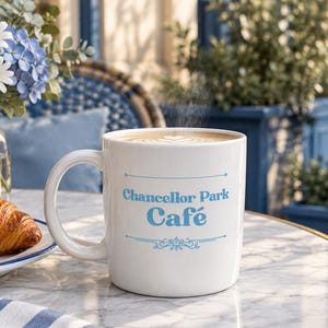 May include: A white ceramic mug featuring the blue text Chancellor Park Café centered between two horizontal lines with decorative flourishes. The mug contains a hot beverage with latte art and sits on a marble table next to a croissant.