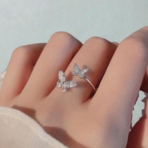 May include: A silver ring with two butterfly motifs, each set with clear, sparkling stones. The ring has an open design, with a butterfly on either side of the finger. The text "BargainNest" is visible at the bottom.