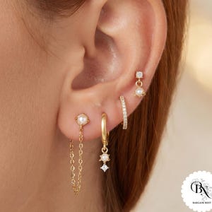 Könnte beinhalten: Ein Set aus vier goldfarbenen Ohrringen, die an einem Ohr getragen werden. Der Schmuck besteht aus einem Perlenstecker mit hängender Kette, einem kleinen Goldring mit Perlen- und Kristallanhänger, einem mit Kristallen besetzten Ring und einem Stecker.