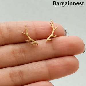 Puede incluir: Un par de pendientes de botón dorados con forma de cuerno. Los pendientes son pequeños y delicados, con un diseño minimalista. El texto "Bargainnest" está en la esquina superior derecha.