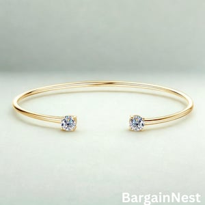 Puede incluir: Una pulsera de brazalete abierta de color dorado con dos piedras preciosas redondas y transparentes en cada extremo. La pulsera tiene un diseño simple y elegante. El texto "BargainNest" es visible en la parte inferior de la imagen.
