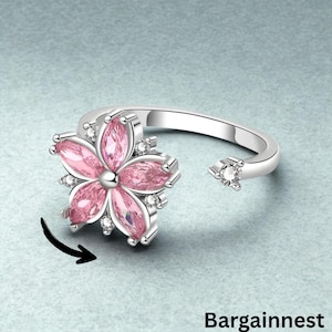May include: A silver-colored ring with a flower design. The flower has pink petals and a central silver-colored bead. Small clear stones are set around the flower and on the open end of the ring. The word "Bargainnest" is at the bottom of the image.