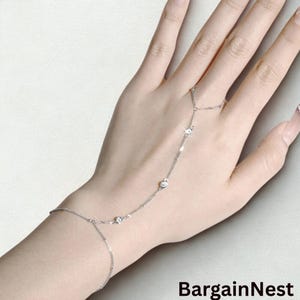 Könnte beinhalten: Ein filigranes silbernes Handkettenarmband mit klaren, runden Edelsteinen. Das Armband verbindet das Handgelenk mit dem Finger mit einer feinen Kette, die mit mehreren funkelnden Steinen besetzt ist. Das BargainNest-Logo ist unten rechts sichtbar.