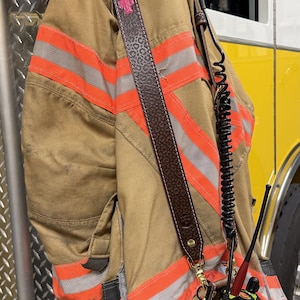 Puede incluir: Una chaqueta de bombero color canela con franjas reflectantes horizontales naranjas y plateadas, que incluye una correa de cuero marrón para radio con textura de leopardo y el nombre J. BURT en letras rosas, unida a una funda con radio.
