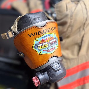 May include: A brown leather fire helmet shield with the text "WIEDECK ESSEX" and a colorful graphic of two fire trucks with flames. The shield is attached to a black and silver fire mask with the "MSA" logo.
