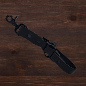 May include: A black leather strap with a metal clasp and buckle. The strap features a swivel hook at one end and a buckle with a loop. The strap is secured with two rivets. The background is a dark wood grain.