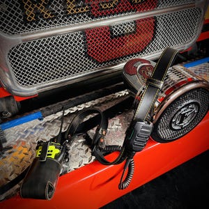 May include: A black leather radio holster and strap set with yellow stitching, featuring a neon yellow two-way radio with an antenna and a black coiled microphone cord. The equipment is displayed against a metallic silver and red vehicle grille.