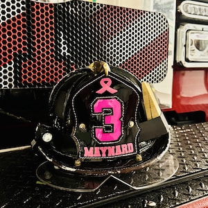 Puede incluir: Un casco de bombero negro con un escudo frontal que presenta un lazo rosa de concienciación sobre el cáncer de mama, el número 3 en grandes dígitos rosas y el nombre MAYNARD en letras mayúsculas rosas, todo bordeado con costuras blancas.