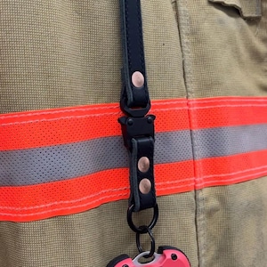 May include: A black leather strap with copper rivets and a black clasp is attached to a pink and black device with the word "Seek" on it. The device is attached to a firefighter's uniform.