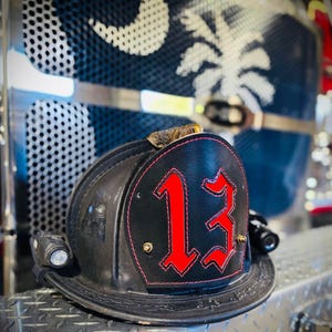 May include: Black firefighter helmet with the number 13 in red on a black leather patch. The helmet has a light attached and is sitting on a diamond-plate surface. The background shows a blue and white design.