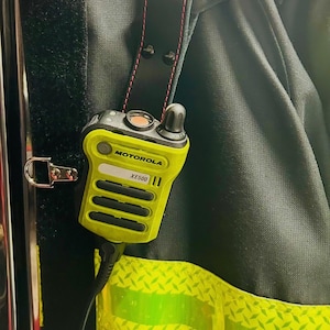 Puede incluir: Una radio Motorola XE500 amarilla y negra sujeta a una correa negra con costuras rojas. La radio está sujeta a una prenda oscura con detalles amarillos reflectantes. La imagen muestra un primer plano de la radio y sus fijaciones.