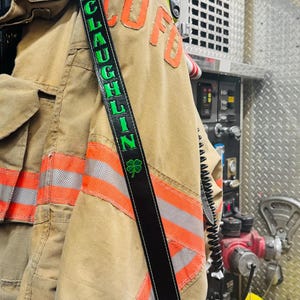 May include: A black leather radio strap draped over a tan firefighter turnout coat with orange and silver reflective stripes. The strap features the name MCLAUGHLIN in bright green lettering, a small green shamrock icon, and a metal rivet.