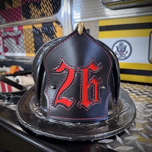 May include: A black leather firefighter helmet front piece featuring the number 26 in bold red lettering with a black outline. The shield is accented with red stitching along the edges and is secured with two silver-toned metal rivets.