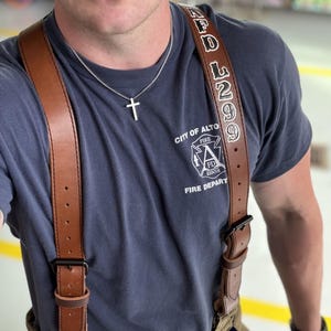 May include: A dark blue t-shirt with the City of Altoona Fire Department logo, a silver cross necklace, and brown leather suspenders featuring the text AFD 299. The suspenders have black metal buckles and are attached to tan trousers.