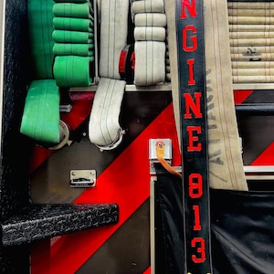 Pode incluir: Um compartimento de caminhão de bombeiros contendo mangueiras dobradas verdes e brancas. Uma alça de couro preta com o texto vermelho ENGINE 813 pende verticalmente sobre uma mangueira bege. O compartimento possui faixas de segurança.