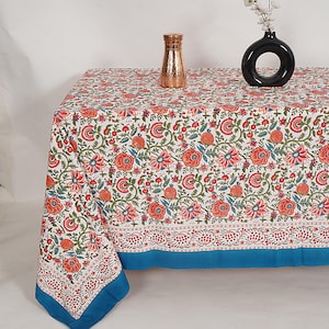 Puede incluir: Un mantel rectangular blanco con un estampado floral en rojo, naranja y verde. El mantel tiene un borde azul y está decorado con un jarrón color cobre y un jarrón negro con flores blancas.