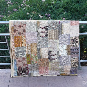 Peut inclure: Une courtepointe patchwork composée de divers tissus à motifs dans des tons beige, marron et crème. De forme rectangulaire, la courtepointe semble faite à la main, avec des coutures visibles. En arrière-plan, une balustrade métallique et un feuillage vert.