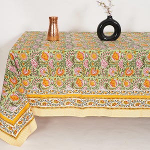 May include: A green floral tablecloth with a repeating pattern of orange and pink flowers and green leaves. The tablecloth has a yellow border and is draped over a table. A copper vase and a black vase with white flowers are on the table.