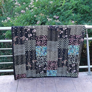Op de afbeelding: Een zwarte patchworkquilt met diverse patronen, waaronder bloemen, chevrons en stippen. De quilt wordt buiten tentoongesteld, over een metalen reling gedrapeerd, met een achtergrond van groen gebladerte en roze bloemen.
