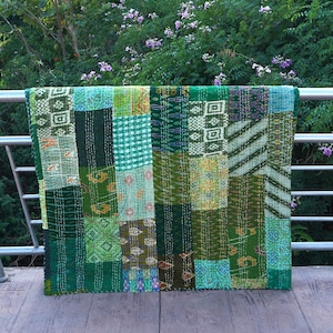 May include: A patchwork quilt with a variety of green fabric squares. The quilt is draped over a metal railing. The quilt features a mix of patterns and textures, including geometric and floral designs. The stitching is visible, adding to the handmade aesthetic.