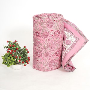May include: A rolled-up pink floral quilt with a matching unfolded section. The quilt features a repeating pattern of pink flowers and green leaves. The border has a striped design. A small potted plant with red berries is on the left.