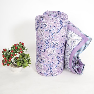 Op de afbeelding: Gerolde lavendelkleurige quilt met een bloemenpatroon in wit en blauw. De quilt is gedeeltelijk uitgerold en onthult een bijpassende rand met een gestreept ontwerp. Een kleine potplant met rode bessen staat op de voorgrond.