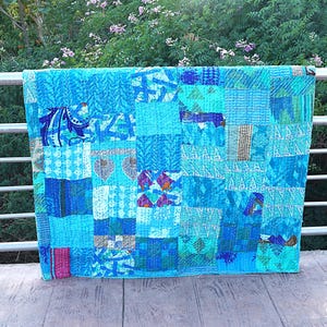 Op de afbeelding: Een patchworkquilt in tinten blauw, blauwgroen en turquoise, met een verscheidenheid aan patronen. De quilt is gedrapeerd over een metalen reling, met een achtergrond van groen gebladerte en paarse bloemen.