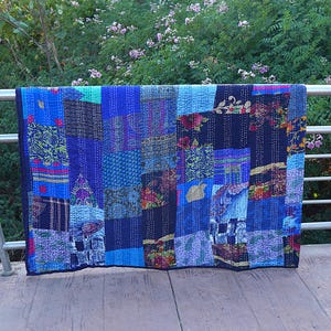 May include: A patchwork quilt in shades of blue, featuring diverse patterns and textures. The quilt is constructed from rectangular fabric pieces, sewn together with white thread. It is draped over a metal railing.