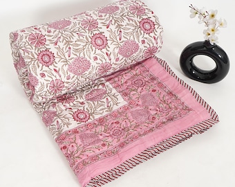 May include: A rolled-up floral quilt with a pink and white pattern. The quilt features a repeating design of pink flowers and green leaves on a white background. The border is pink with a striped edge. A black vase with white flowers is in the background.