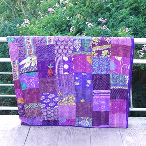 Könnte beinhalten: Ein Patchwork-Quilt in Lilatönen, im Freien ausgestellt. Der Quilt besteht aus verschiedenen Stoffquadraten mit unterschiedlichen Mustern und Texturen, darunter Blumen- und geometrische Designs. Der Quilt ist über einem weißen Geländer vor dem Hintergrund grüner Blätter drapiert.