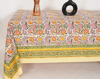 May include: A white tablecloth with a floral pattern in pink, yellow, and green. The tablecloth has a border of green and yellow floral designs. A copper vase and a black vase with a circular opening are on the table.