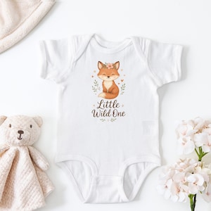 May include: White baby onesie with a watercolor illustration of a fox wearing a floral crown. The text "Little Wild One" is below the fox. A stuffed teddy bear and a blanket are next to the onesie.