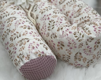 Pode incluir: Um ninho de bebê com um padrão floral em tons de marrom e rosa sobre um fundo branco. As extremidades são feitas de tecido mauve texturizado. Uma flor branca e uma corda acentuam o design.
