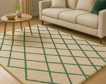 Op de afbeelding: Een beige vloerkleed met een groen ruitpatroon. Het kleed ligt op een lichte houten vloer voor een beige bank. Een kleine houten bijzettafel en een raam zijn ook zichtbaar op de achtergrond.