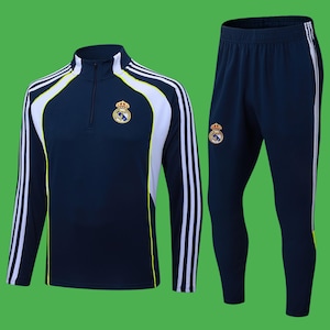 May include: Navy blue athletic tracksuit featuring a long-sleeved top and matching pants. The top has a quarter-zip collar, white and yellow accents, and the team logo on the chest. The pants have white stripes down the side and the team logo.
