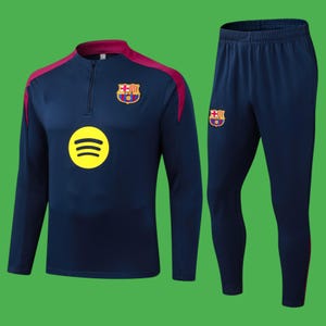 Puede incluir: Un chándal azul marino, compuesto por una parte superior de manga larga y pantalones a juego. La parte superior tiene cuello y detalles en los hombros burdeos, cierre de cremallera y un logotipo amarillo de Spotify. Ambas piezas muestran un escudo del equipo.