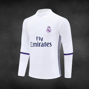 K&ouml;nnte beinhalten: Wei&szlig;es Langarmtrikot mit Stehkragen. Das Trikot tr&auml;gt den Text "Fly Emirates" in Marineblau sowie ein Teamwappen. Marineblaue und violette Akzente befinden sich an den &Auml;rmeln.