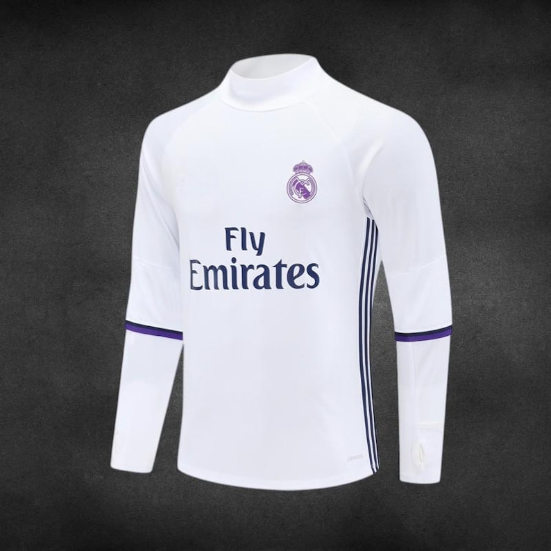 K&ouml;nnte beinhalten: Wei&szlig;es Langarmtrikot mit Stehkragen. Das Trikot tr&auml;gt den Text "Fly Emirates" in Marineblau sowie ein Teamwappen. Marineblaue und violette Akzente befinden sich an den &Auml;rmeln.
