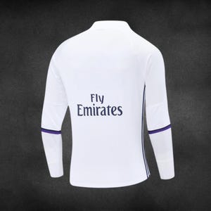 K&ouml;nnte beinhalten: Wei&szlig;es Langarm-Sportshirt mit lila Akzenten. Der Schriftzug "Fly Emirates" ist in Marineblau gehalten. Lila B&auml;nder befinden sich an den Handgelenken, mit einem vertikalen Streifen an der Seite.