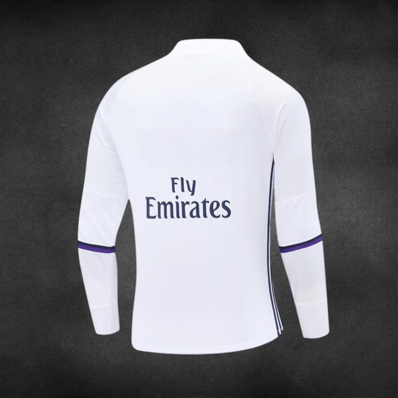 K&ouml;nnte beinhalten: Wei&szlig;es Langarm-Sportshirt mit lila Akzenten. Der Schriftzug "Fly Emirates" ist in Marineblau gehalten. Lila B&auml;nder befinden sich an den Handgelenken, mit einem vertikalen Streifen an der Seite.