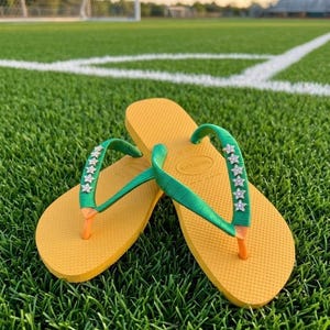 May include: Yellow flip-flops with green straps adorned with star-shaped embellishments. The flip-flops are resting on a green grassy surface, with a soccer field and stadium in the background. The flip-flops have orange toe posts.
