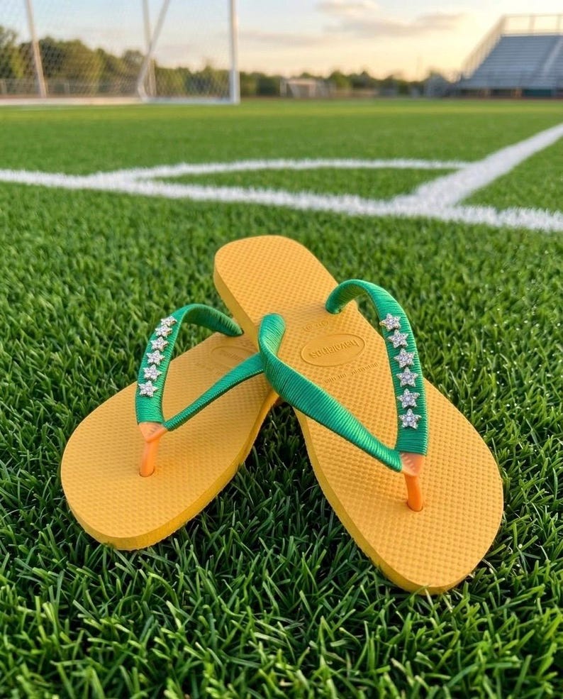 May include: Yellow flip-flops with green straps adorned with star-shaped embellishments. The flip-flops are resting on a green grassy surface, with a soccer field and stadium in the background. The flip-flops have orange toe posts.