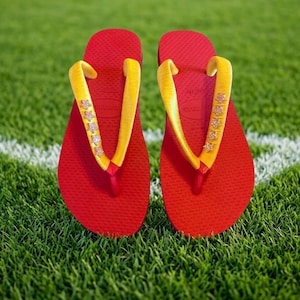 May include: A pair of red flip-flops with yellow straps adorned with star-shaped embellishments. The flip-flops are resting on a bed of green grass, with a white line visible in the background. The sandals are a vibrant summer accessory.