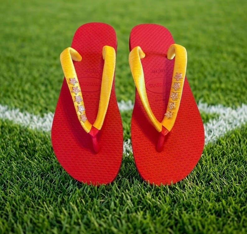 May include: A pair of red flip-flops with yellow straps adorned with star-shaped embellishments. The flip-flops are resting on a bed of green grass, with a white line visible in the background. The sandals are a vibrant summer accessory.