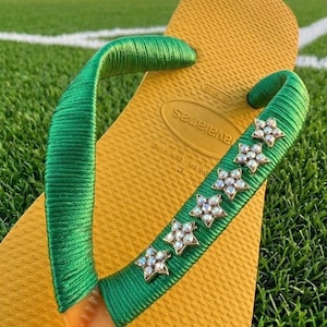 May include: Yellow flip-flop with green straps. The straps are embellished with sparkling star-shaped rhinestones. The flip-flop is resting on a green grassy surface, suggesting outdoor wear.