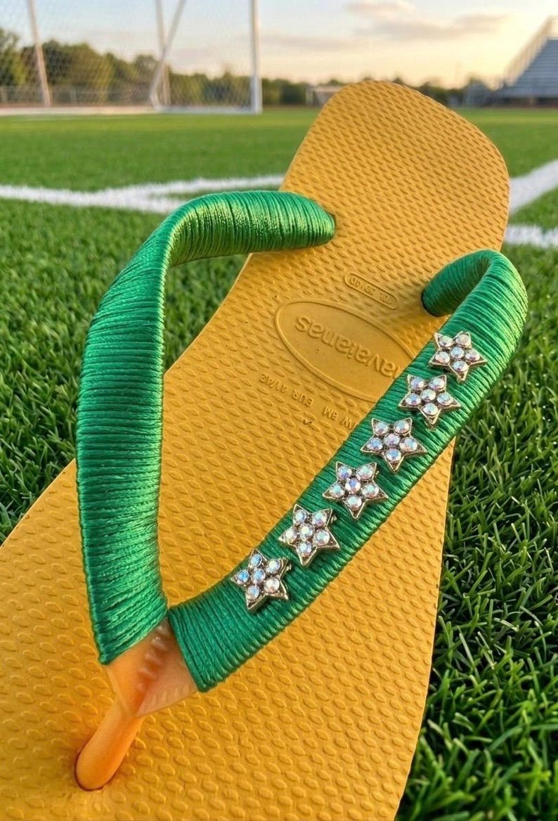 May include: Yellow flip-flop with green straps. The straps are embellished with sparkling star-shaped rhinestones. The flip-flop is resting on a green grassy surface, suggesting outdoor wear.